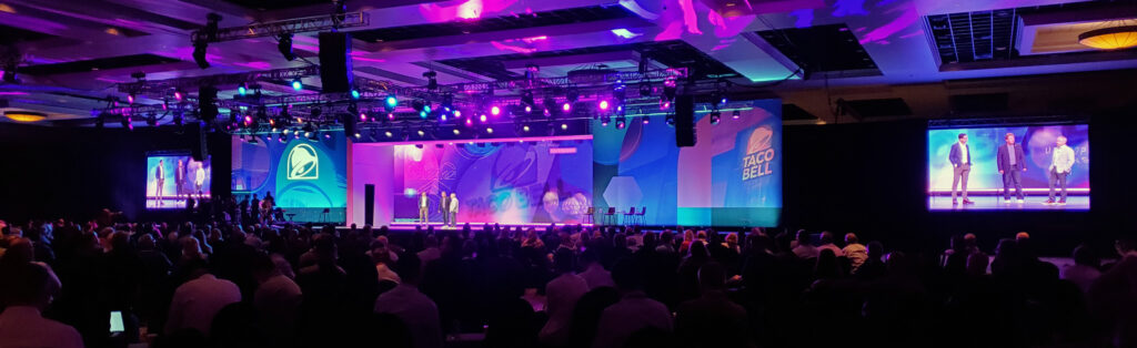 A large audience watches a presentation on stage with purple lighting at a Taco Bell corporate event, with two screens displaying the speakers.